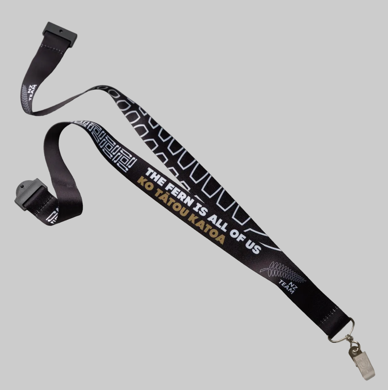 A black fabric lanyard with white graphic patterns and text reading 'THE FERN IS ALL OF US KO TATOU KATOA' in gold, shown on a light gray background with a metal clip at one end.