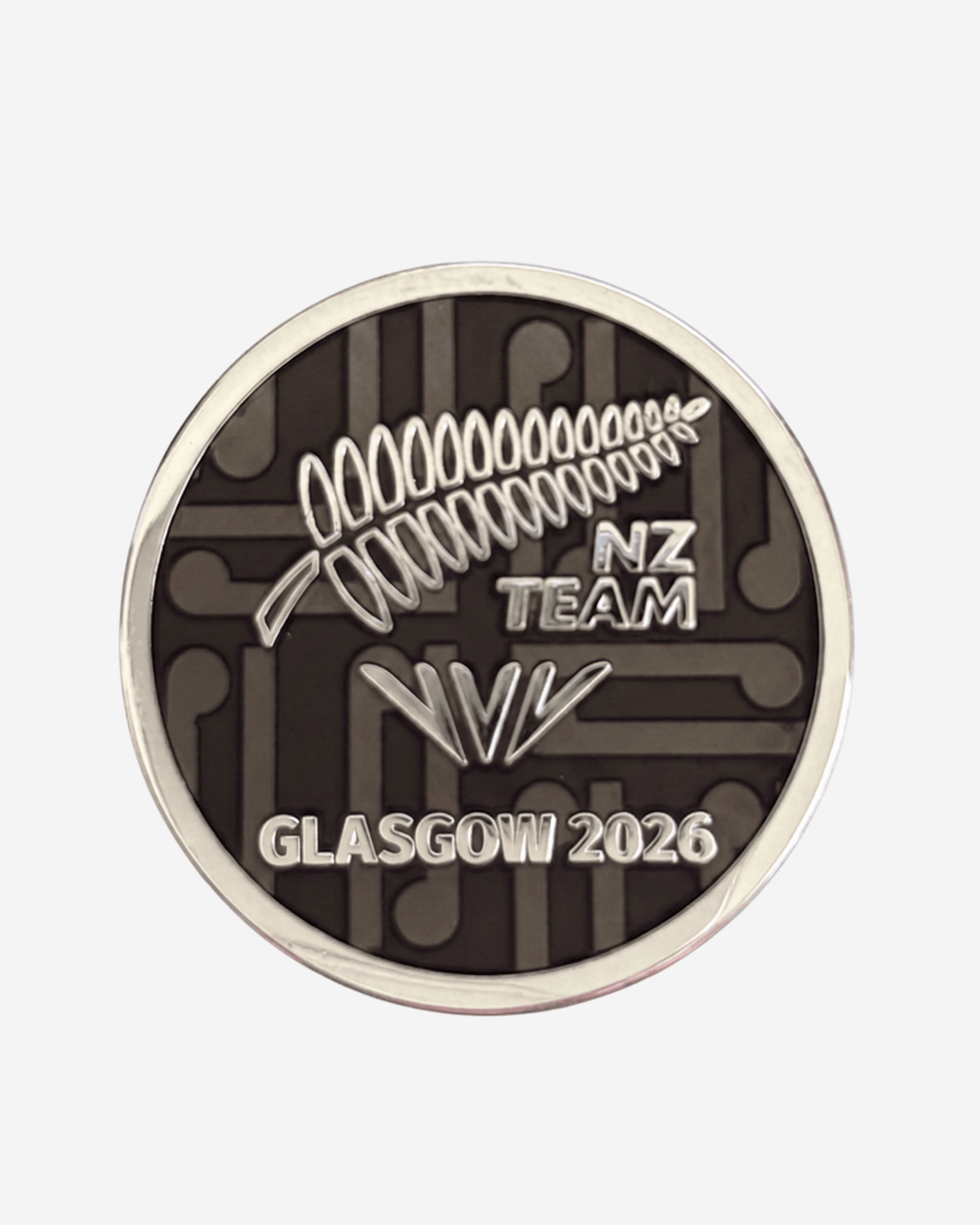 A circular commemorative coin or medallion for the NZ Team at the Glasgow 2026 event, featuring a silver fern and 'NZ TEAM' text on a dark brown background with circuit-like patterns, surrounded by a silver rim.