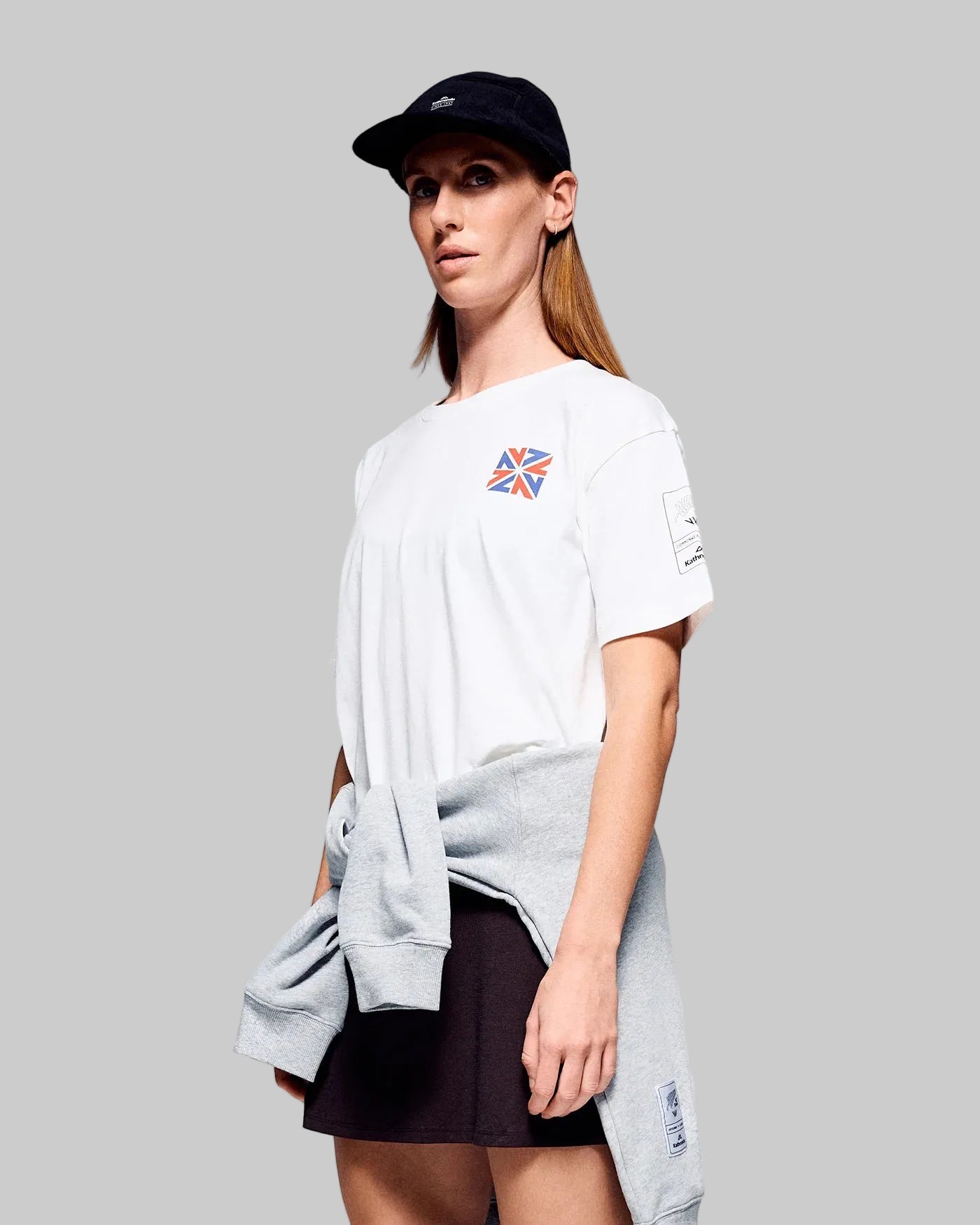 Woman wearing a white t-shirt with a colorful logo, gray sweatshirt, and black cap on a gray background