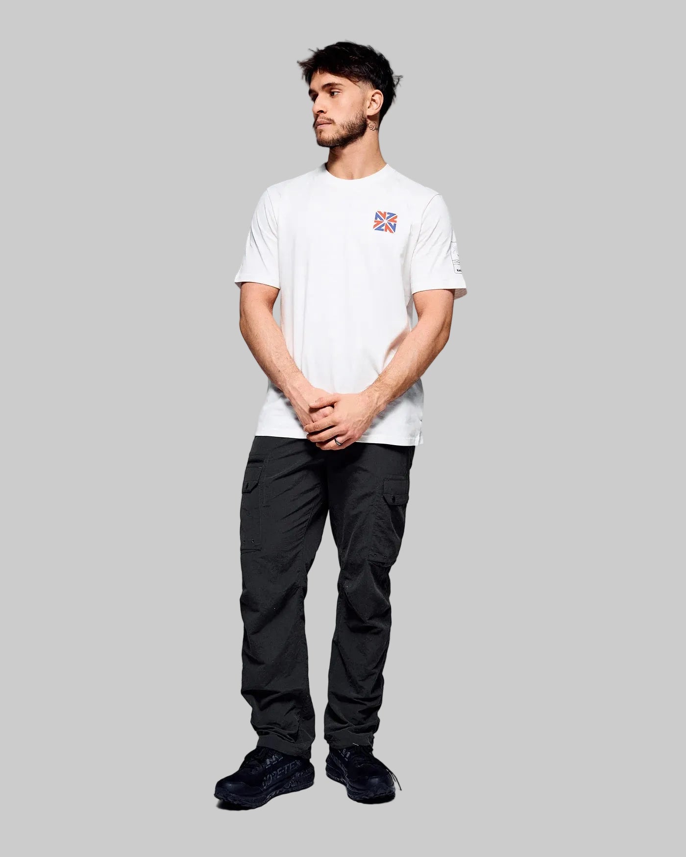 A man modeling a white short-sleeve crewneck t-shirt with a small red and blue graphic on the left chest, paired with black cargo pants and black sneakers, against a light gray studio background.